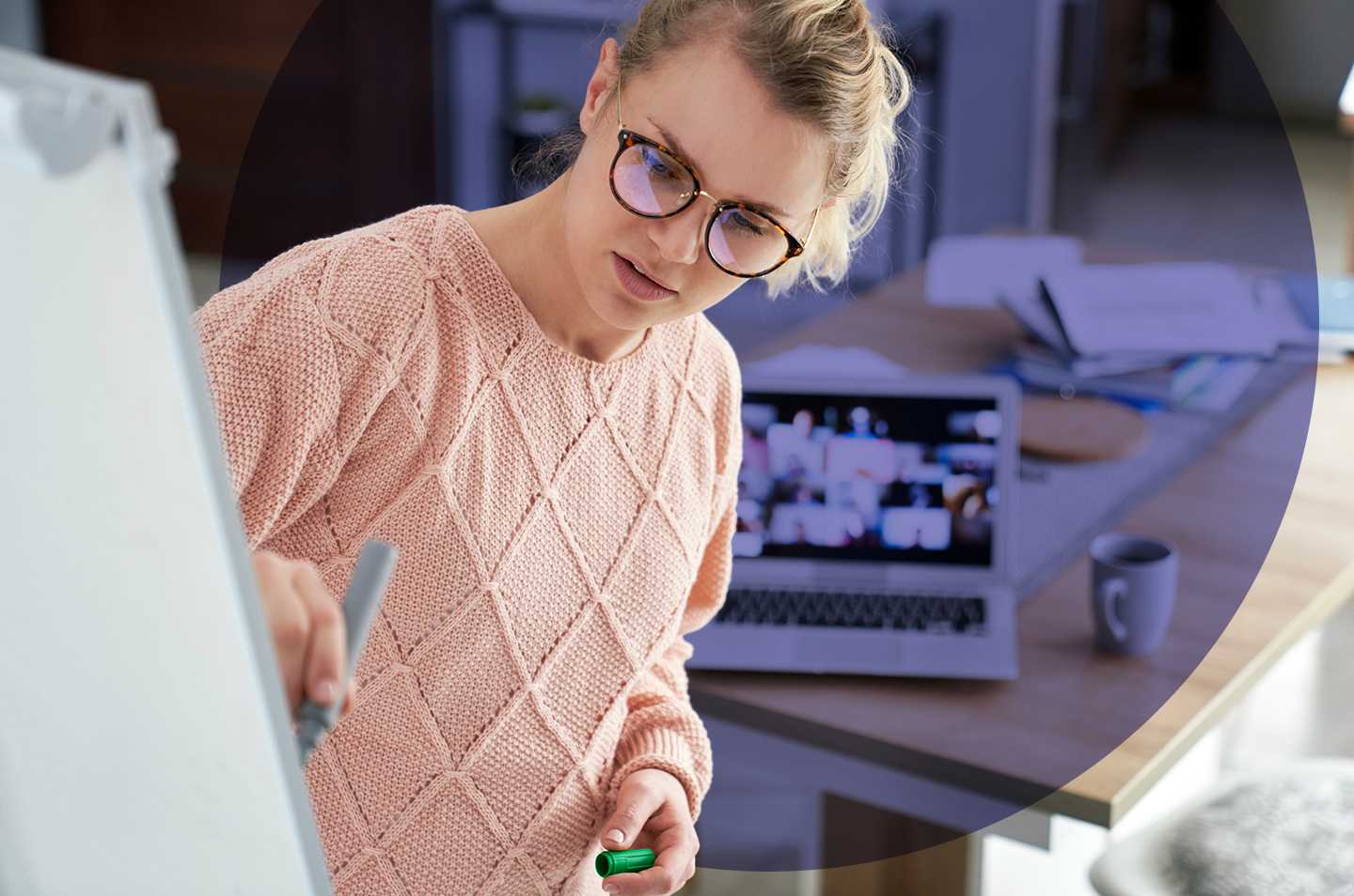 a woman writing on a whiteboard with a laptop on a desk in the background